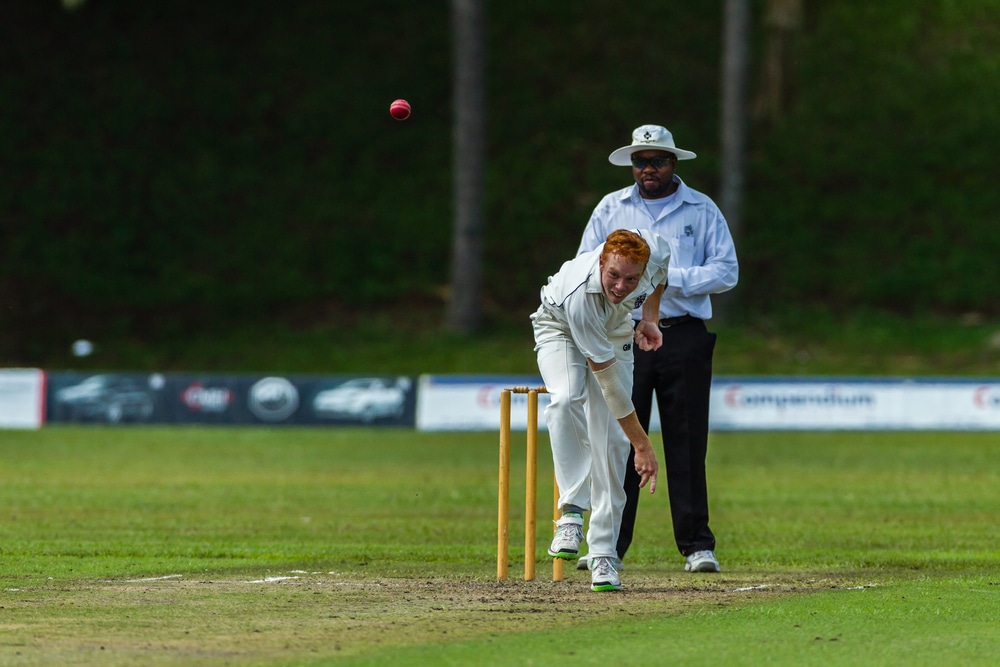 A bowler bowls a red cricket ball. An umpire is seen behind him.