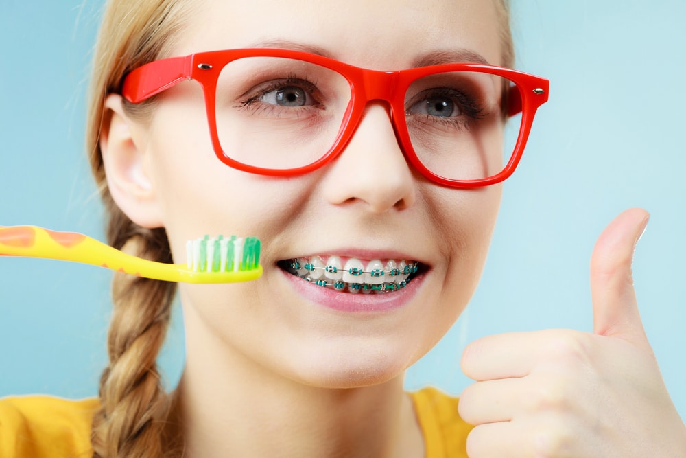 A girls wearing traditional braces holds up a toothbrush and gives a thumbs up.